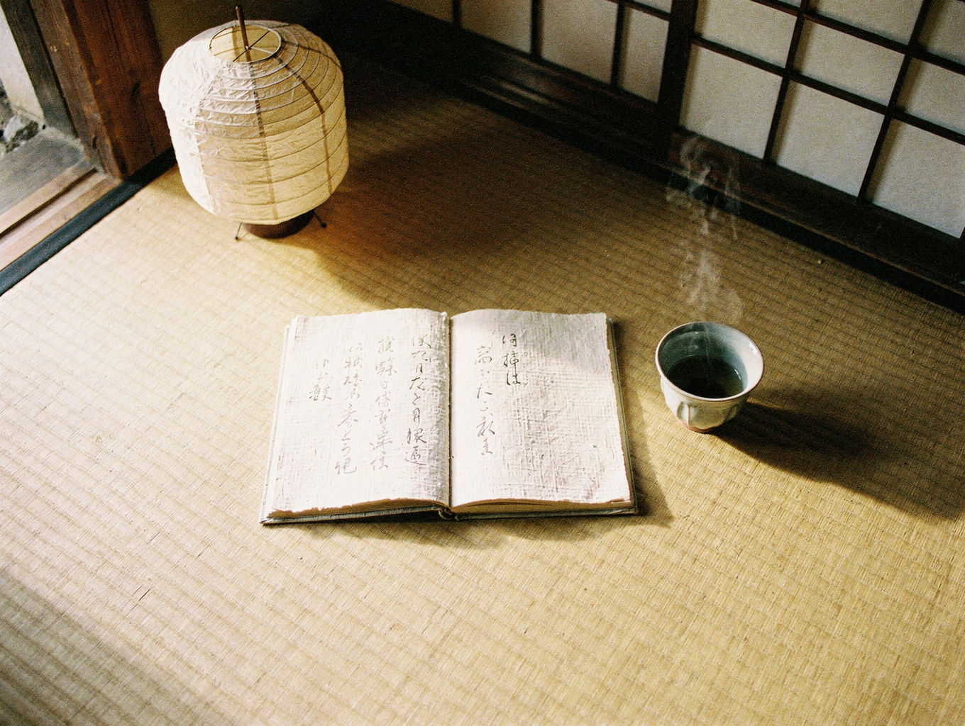 An open notebook of washi, a ceramic cup, and a paper lamp on tatami in morning light.